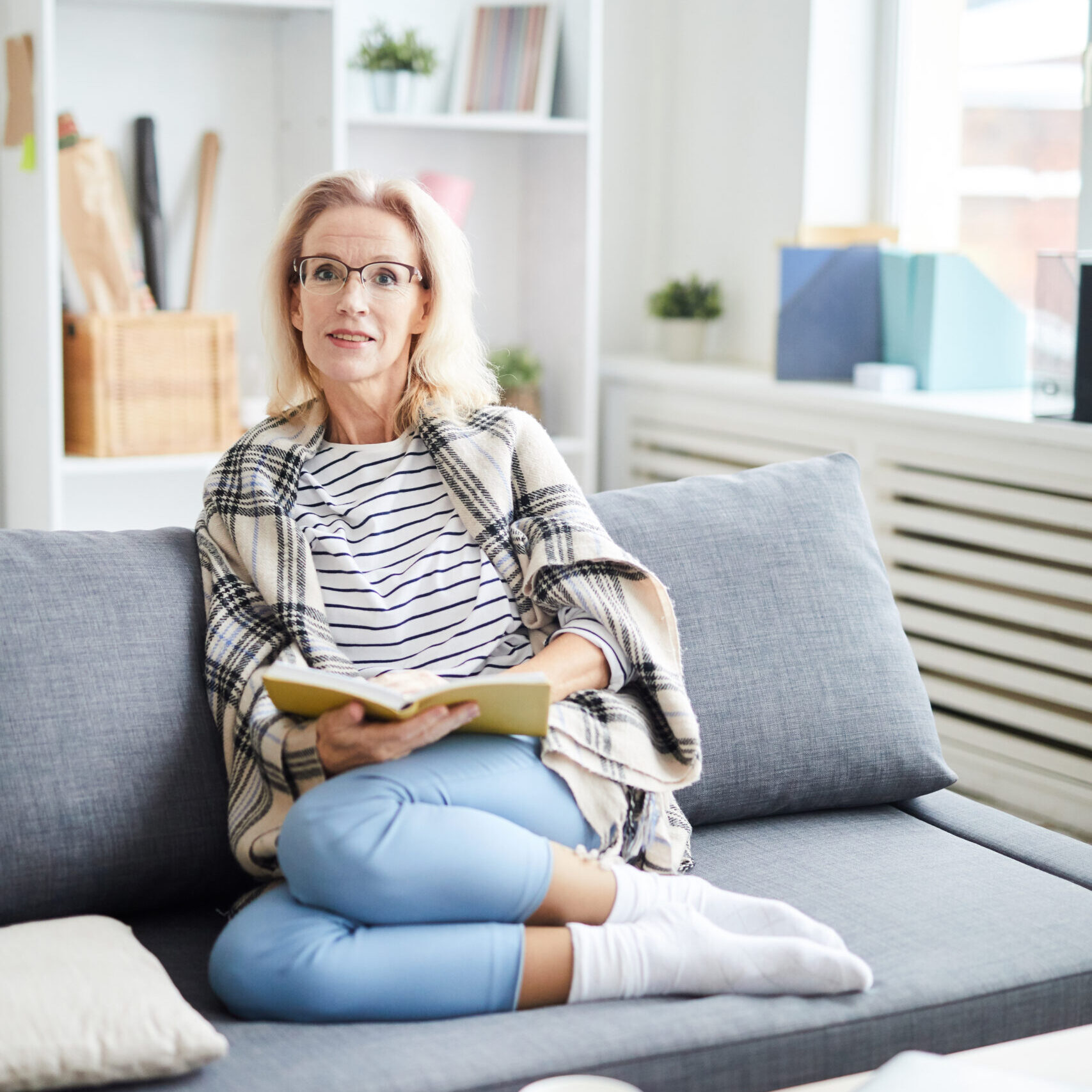 Full length portrait of contemporary woman looking at camera while reading book sitting on sofa at home, copy space