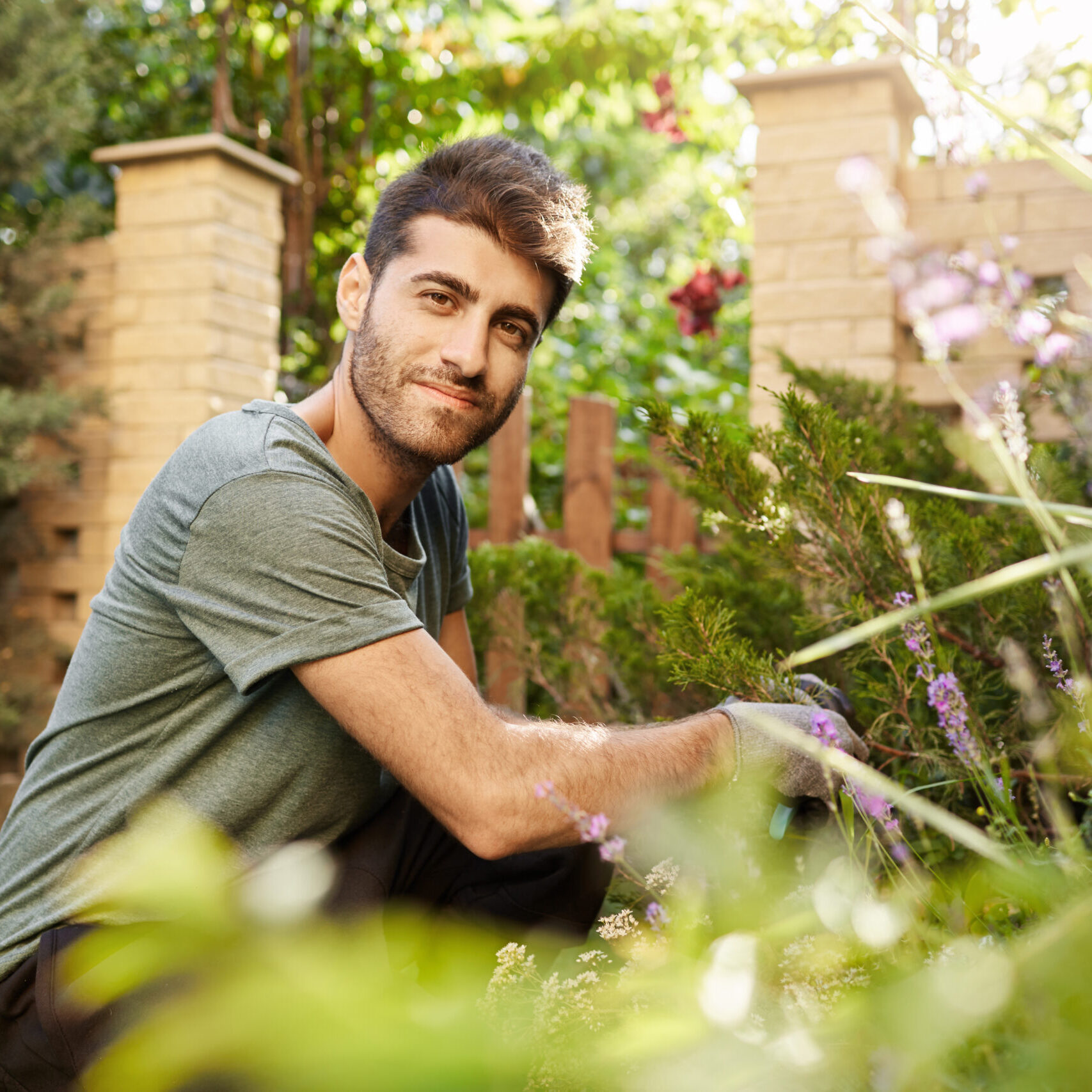 Outdoors portrait of young attractive bearded caucasian man in blue -shirt and sport pants smiling, sitting on grass, looking in camera with happy face expression, working in garden